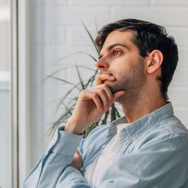 Man in a light blue shirt and white T-shirt gazing into space with his right hand at his mouth - How To Write IELTS General Training Task 1