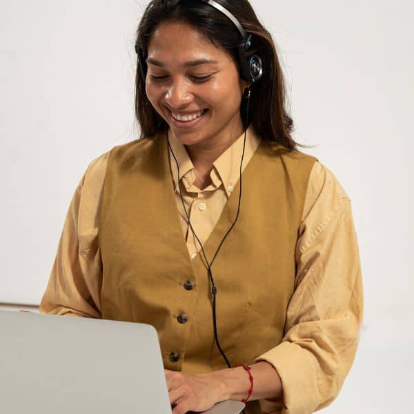 A smiling dark-haired woman in coffee coloured shirt and brown waistcoat typing on a laptop - Tone / Formality for Better Letters & Emails - IELTS