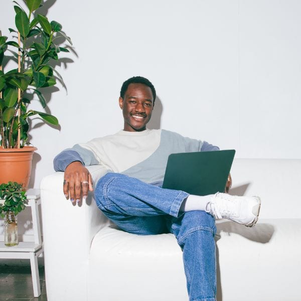 smiling man with short hair sitting with his legs crossed in a white chair beside a plant with a laptop on his knee in a white room - Latest IELTS Writing Task 2 Essays by Topic