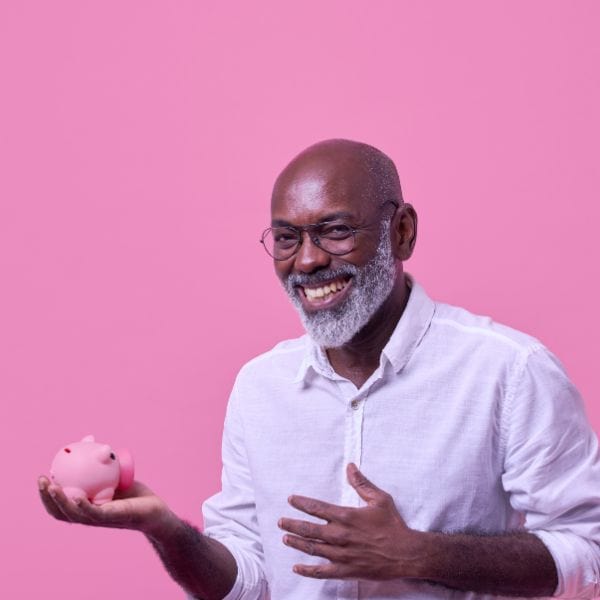 Smiling man with grey beard holding a little pink piggy bank in his right hand in front of a pink background - IELTS Speaking Part 1: 16 Great Ways Start Your Test!