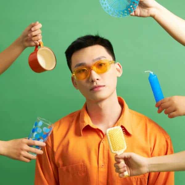 Man with short dark hair, yellow glasses, wearing an orange shirt with other hands holding various items against a green background - IELTS Topic Vocabulary for Writing Task 2